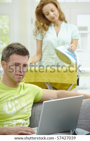 Man with broken leg resting on couch, using laptop computer. Woman doing ironing at the background.