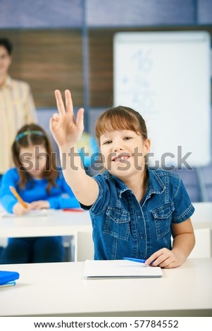 Schoolgirl raising hand to answer question smiling, other girl and teacher in background of class.?