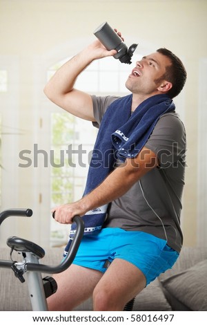 Man sitting on stationary bike at home and drinking water from bottle.