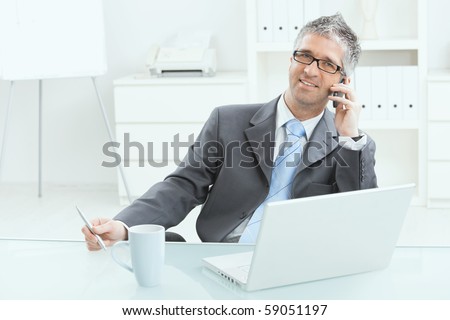 Gray haired executive businessman working on laptop computer at desk, in office, calling on mobile phone.
