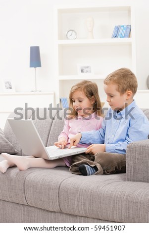 Little brother and sister sitting together on couch at home, using laptop computer, looking at screen.