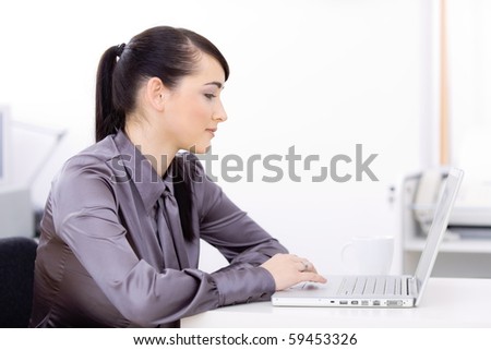Profile portrait of young businesswoman using laptop computer at office desk, looking at screen.