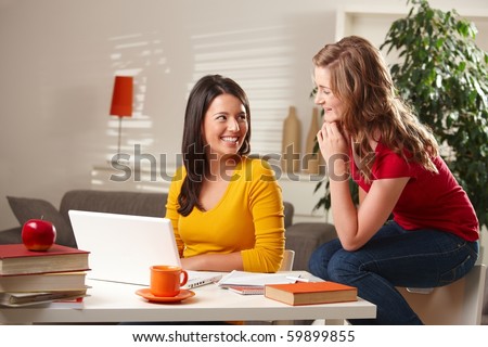 Happy teenage girls laughing together, sitting at table with laptop and books at home.?