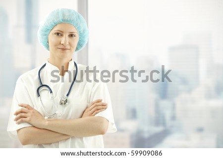 Mid adult female medical doctor standing in skyscraper office with arms folded, wearing white smock.?