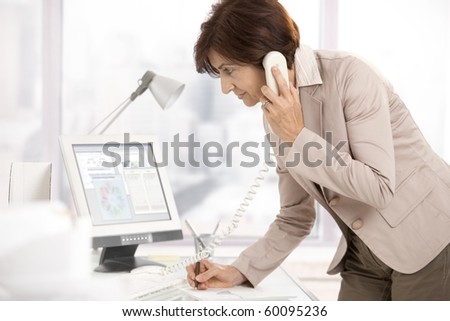 Senior businesswoman at work, standing at desk on landline phone, writing notes.?