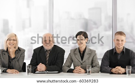 Formal businessteam portrait of different generations sitting at meeting table, smiling at camera.?