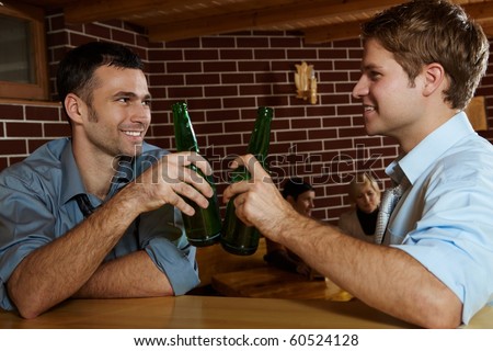 Two men drinking beer in bar, clinking bottles, smiling, women talking in background.?
