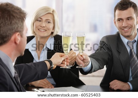Happy young business people sitting around meeting table at office celebrating success with champagne, smiling. Focus placed on flutes.