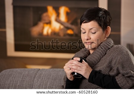 Woman sitting on sofa at home on a cold winter day, drinking hot tea, looking down.