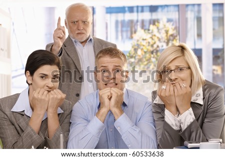 Scared employees sitting with hands up to mouth, angry boss standing behind pointing.?