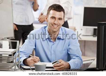 Casual young businessman working in office, sitting at desk, writing notes to personal organizer.