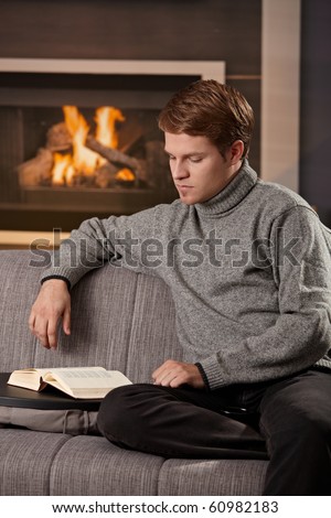 Young man sitting on sofa at home on a cold winter day, reading book in front of fireplace.
