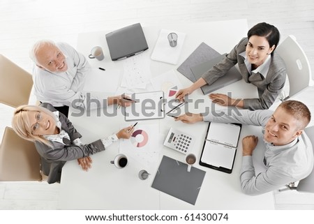 Smiling businesspeople sitting at meeting table, working, pointing at document, smiling at camera, high angle view.