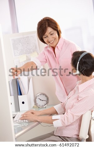 Smiling supervisor pointing, helping customer service operator sitting at desk.?