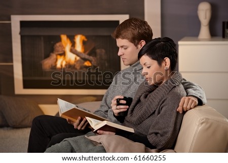 Young couple hugging on sofa in front of fireplace at home, reading books.