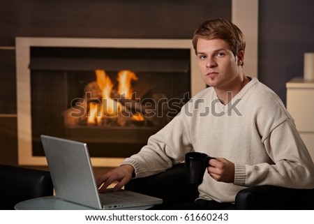 Young man sitting in front of fireplace at home on a cold winter day, working on laptop computer.