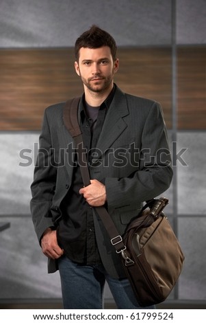 Handsome trendy office worker standing in lobby holding laptop bag looking at camera confidently.
