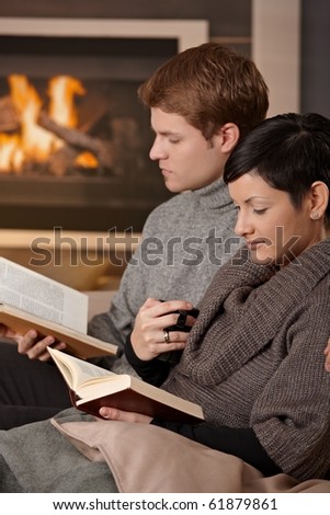 Young couple sitting on sofa in front of fireplace at home, reading books.