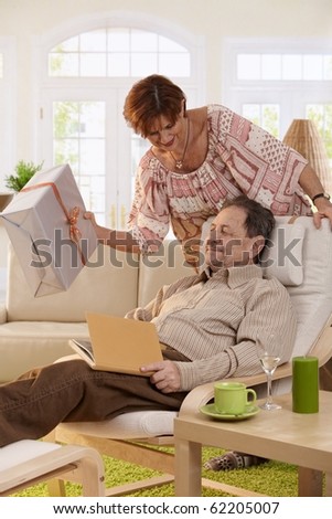 Senior couple celebrating birthday. Wife giving present to her husband, resting in armchair.?