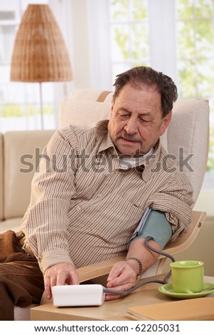 Old man sitting in armchair at home, measuring his blood pressure. ?