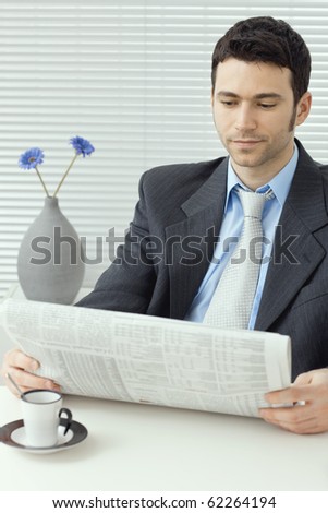 Young businessman having a morning coffee break, sitting at desk and reading business news.