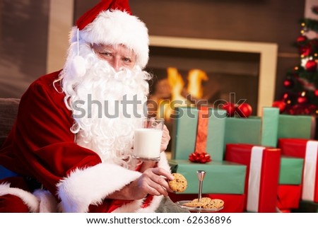 Happy Santa Claus sitting at fireplace drinking milk and eating chocolate chip cookies, looking at camera.?
