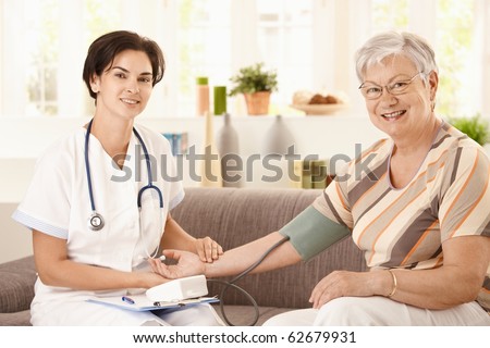 Nurse measuring blood pressure of senior woman at home. Looking at camera, smiling.?