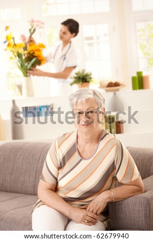 Portrait of happy senior woman sitting on sofa at home, nurse arranging flowers in background.?