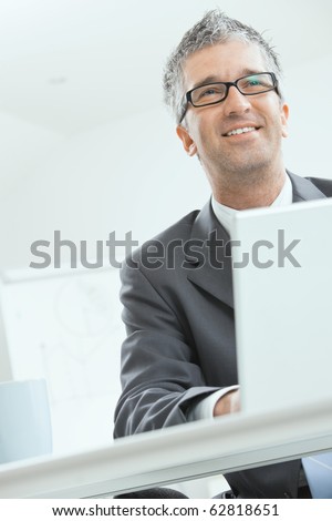 Businessman sitting at desk and working with laptop computer. Looking at camera, smiling.