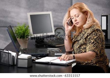 Senior woman sitting at office desk talking on mobile phone matching dates from personal organizer.?