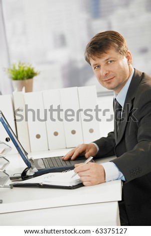 Businessman working in office, writing notes to organizer and using laptop computer.?