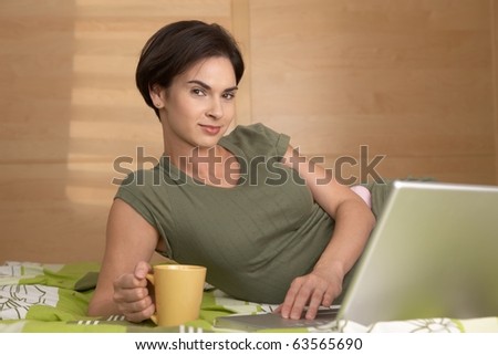 Morning portrait of smiling woman looking at camera, holding coffee mug and using laptop lying on bed.?