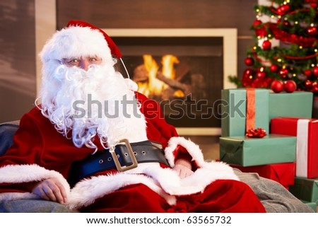 Portrait of Santa Claus sitting by fireplace with Christmas presents all around, looking at camera.?