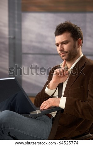 Troubled young man working on laptop computer, trying to solve problem.