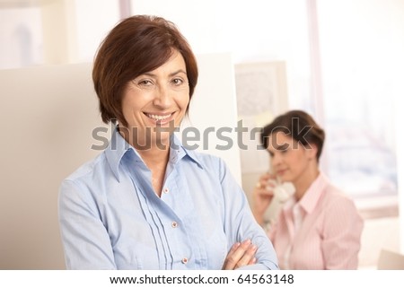 Senior professional woman smiling at camera, assistant using land line phone in background.