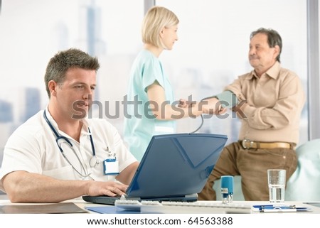 Mid-adult doctor sitting at desk looking at computer, nurse measuring blood pressure of older patient in office.