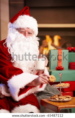 Happy Santa Claus sitting at fireplace drinking milk eating chocolate chip cookies, looking at camera, winking?