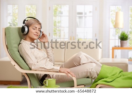 Woman relaxing in armchair at home enjoying music in headphones, smiling.?
