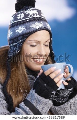 Pretty young girl dressed up warm for skiing wearing cap and gloves drinking hot drink eyes closed front of winter landscape, smiling.?