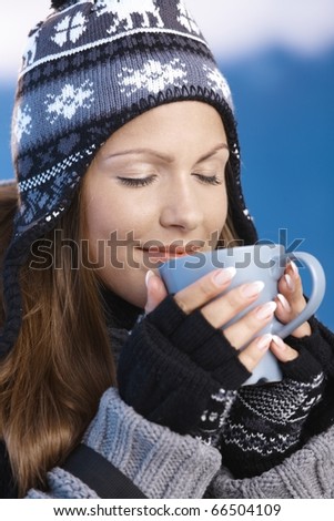 Pretty young girl dressed up warm for skiing wearing cap and gloves drinking hot drink eyes closed front of winter landscape .?