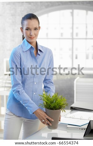 Young businesswoman holding potted plant standing at office desk, smiling at camera.?
