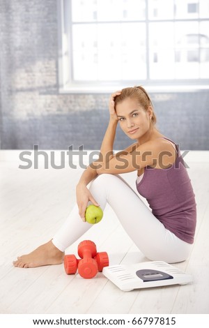 Portrait of pretty sporty girl sitting on floor in gym, holding green apple, with scale and dumbbells, smiling at camera.?