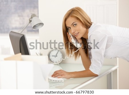 Portrait of working office girl at desk, using landline phone and desktop computer, smiling happily at camera.?