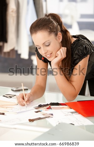 Young, attractive fashion designer working in office, drawing, leaning on desk, smiling.?