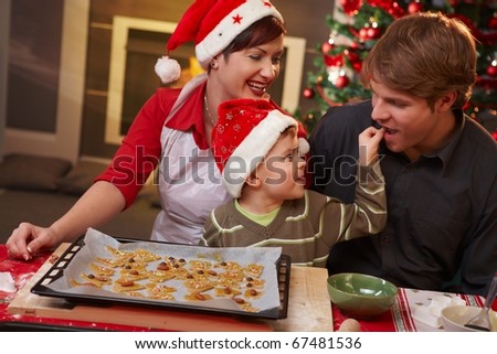 Small son helping dad tasting christmas cake at table, mother watching, laughing.