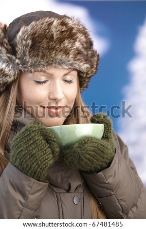 Young attractive female dressed up warm in coat, fur-hat and gloves, drinking hot tea, smiling, eyes closed.