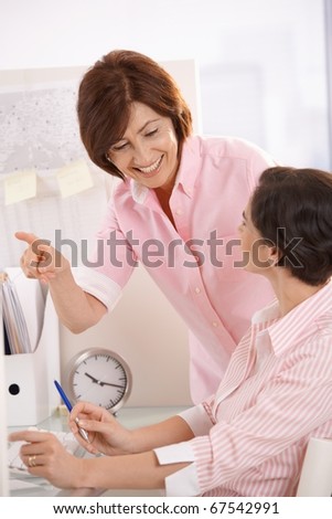 Senior office worker teaching coworker in office, smiling, pointing.?