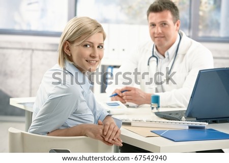 Smiling woman sitting in doctor\'s office on appointment, looking at camera, doctor in background.?