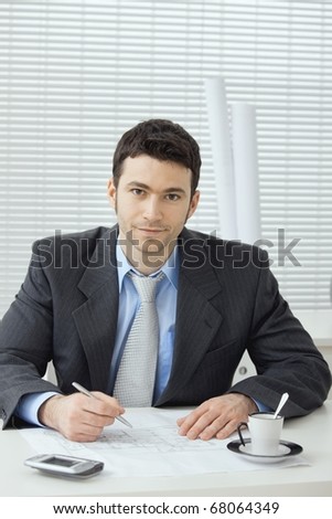 Architect wearing grey suit sitting at office desk, writing notes on floor plan. Looking at camera, smiling.?