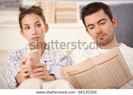 Young couple in bed, man reading newspaper, woman bored, drinking tea.